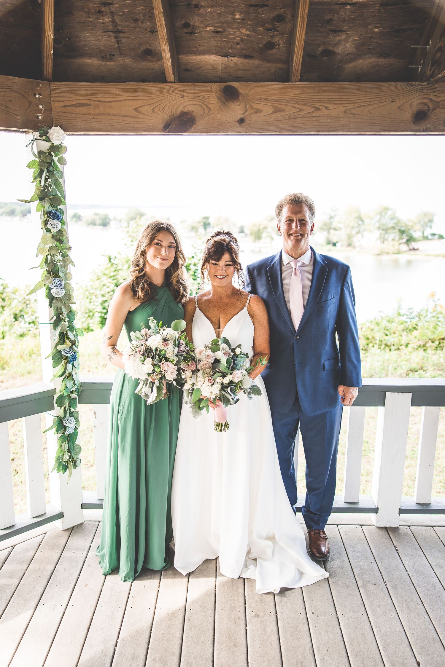 Wedding party in green dresses and blue suit poses on a wooden deck with greenery decorations.