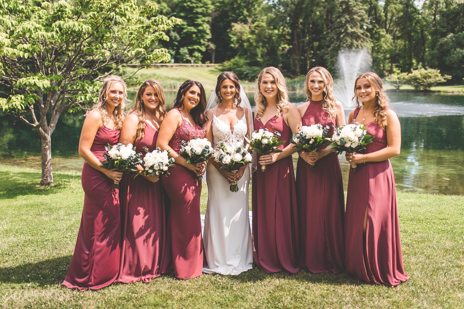 Wedding party members in burgundy dresses pose together on grass with white floral bouquets.