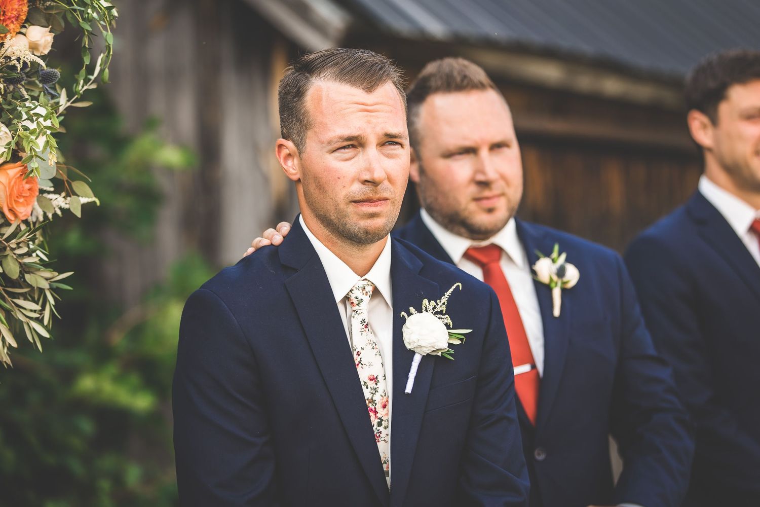 Wedding party members in navy suits with floral ties await during ceremony.