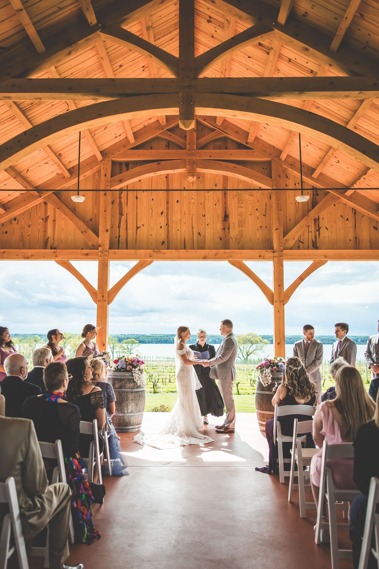 Wedding ceremony under wooden beams of an open-air pavilion overlooking scenic countryside.