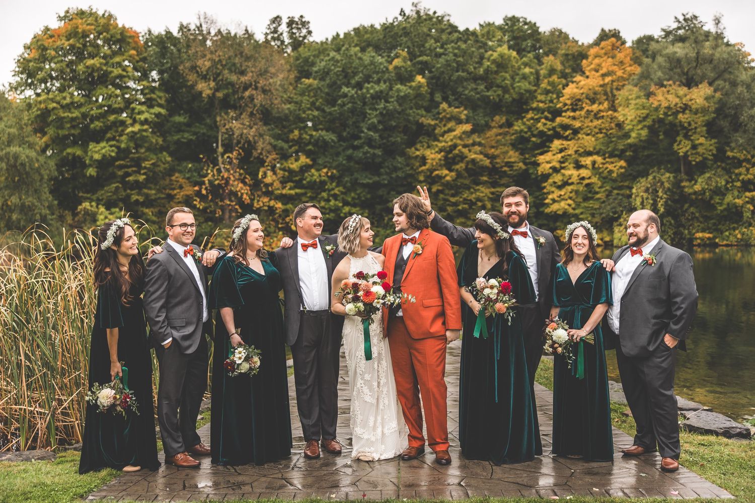 Wedding group photo with bridal party in black attire against fall foliage backdrop.