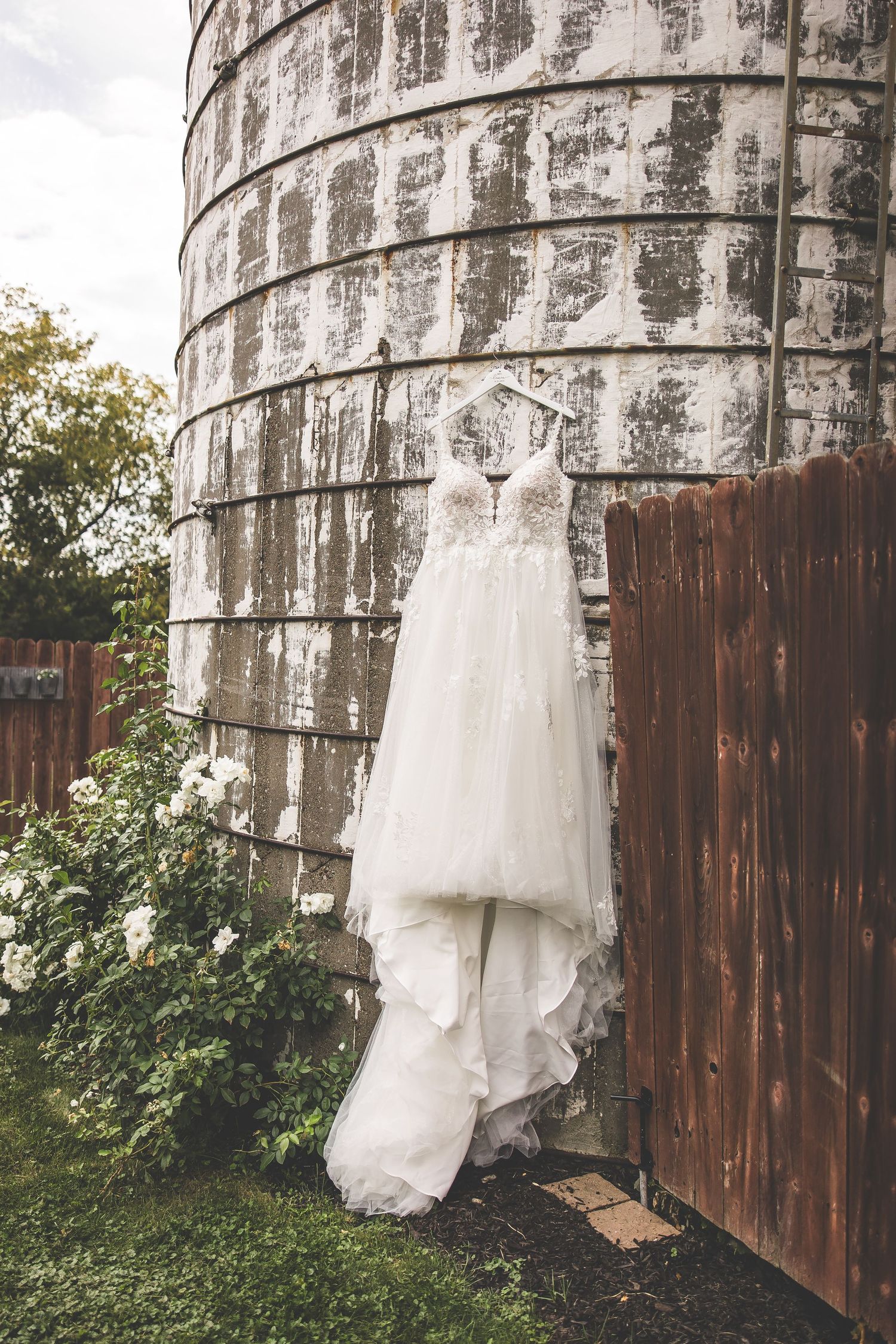 White wedding dress hanging against a weathered silo wall with wooden fence in background.