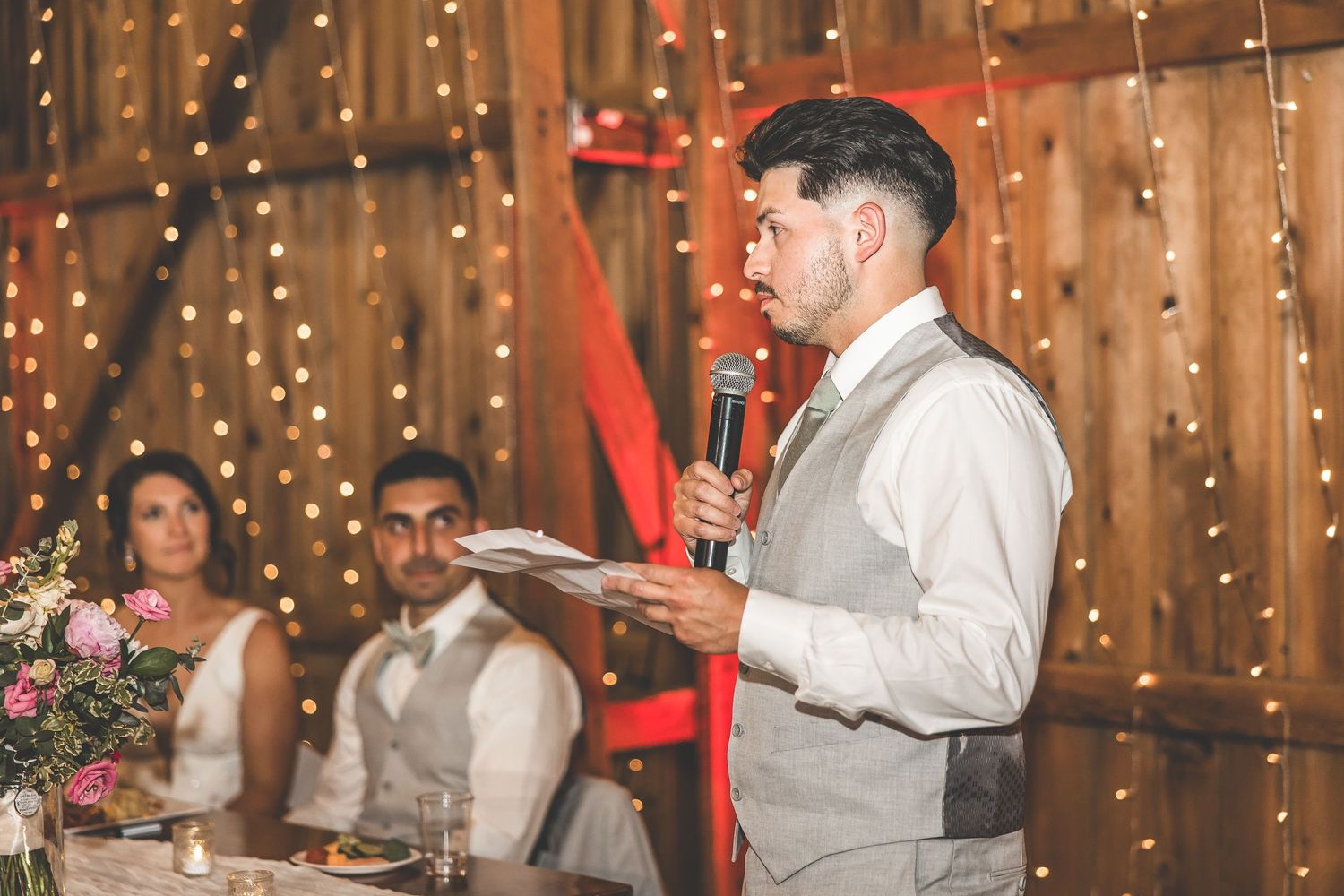 Wedding guests dance and celebrate in a rustic barn venue decorated with string lights and floral arrangements.