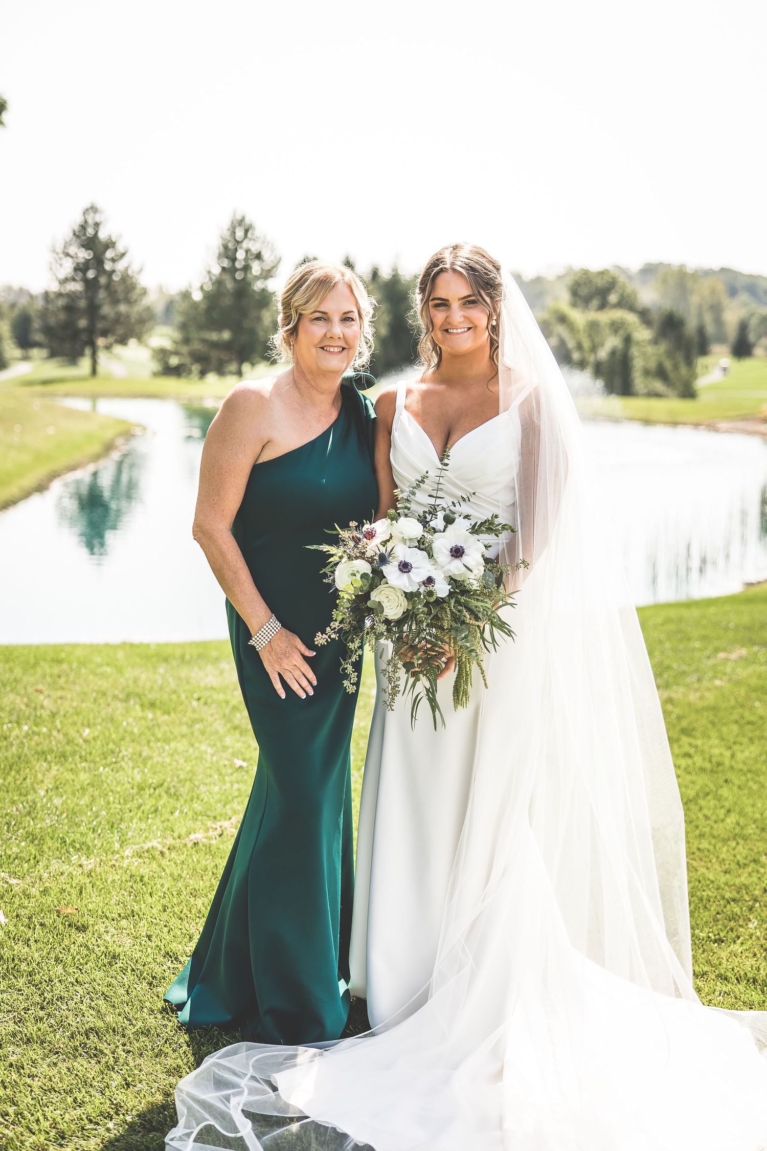 Wedding party poses on grassy hill with lake view in sage green and white dresses.