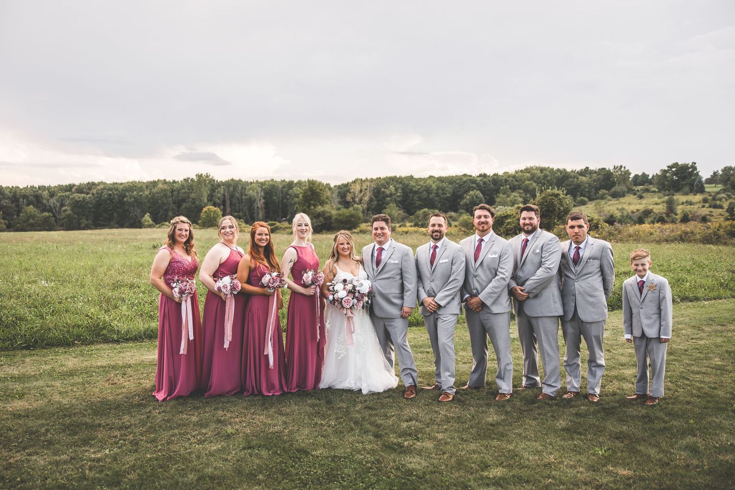 Wedding party members lined up in pink dresses and gray suits for formal outdoor portrait.