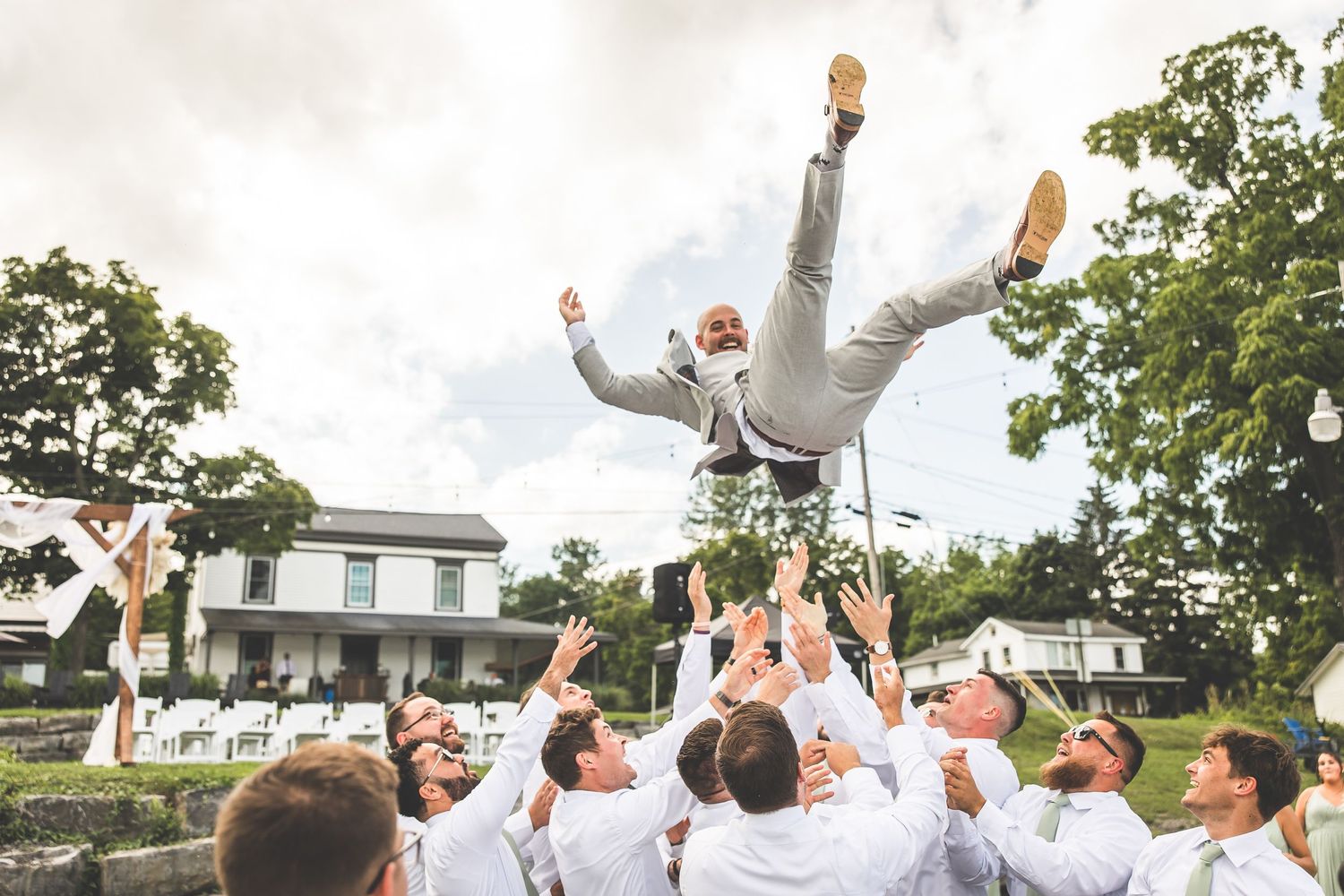 Groomsmen toss someone in gray suit into air during outdoor wedding celebration.