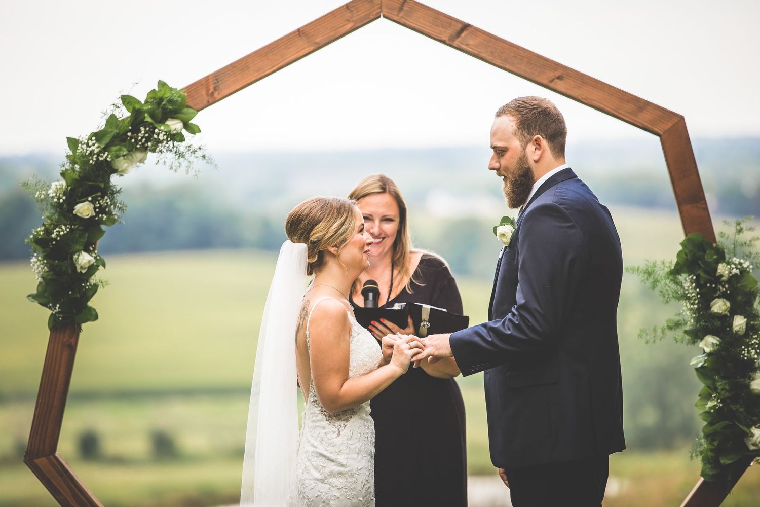 Wedding ceremony under floral-adorned hexagonal arch with rolling hills in background.