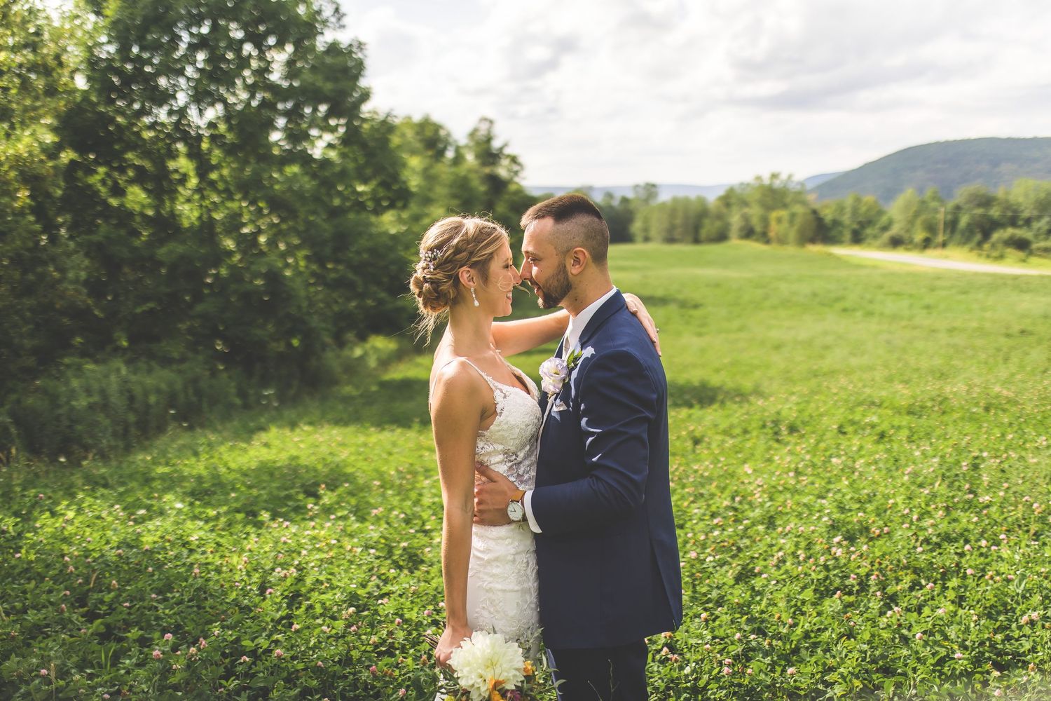 Wedding couple poses in a scenic field with mountains and trees visible in the background.