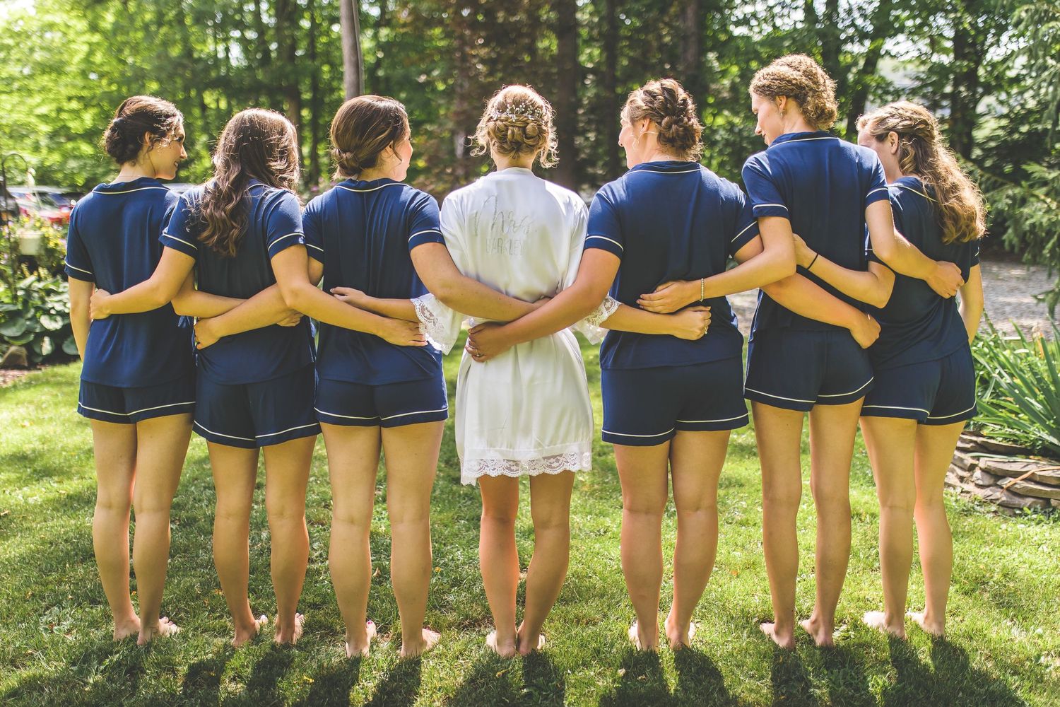 Back view of bridesmaids in navy robes standing with arms around each other in garden setting.
