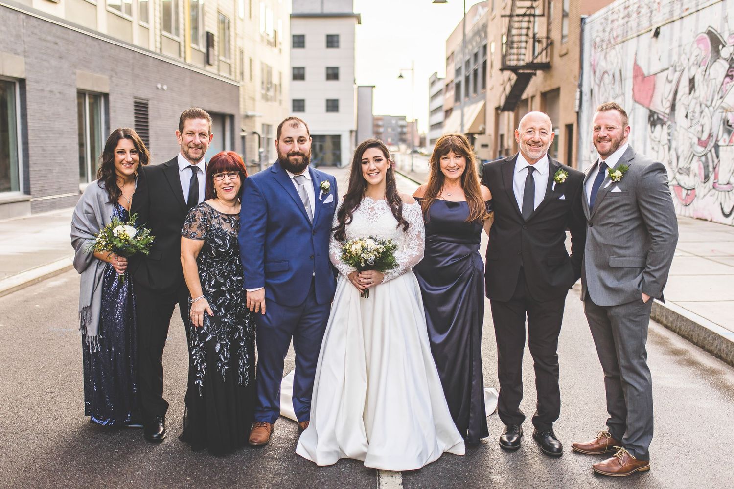 Large wedding party poses together on urban street with brick buildings.