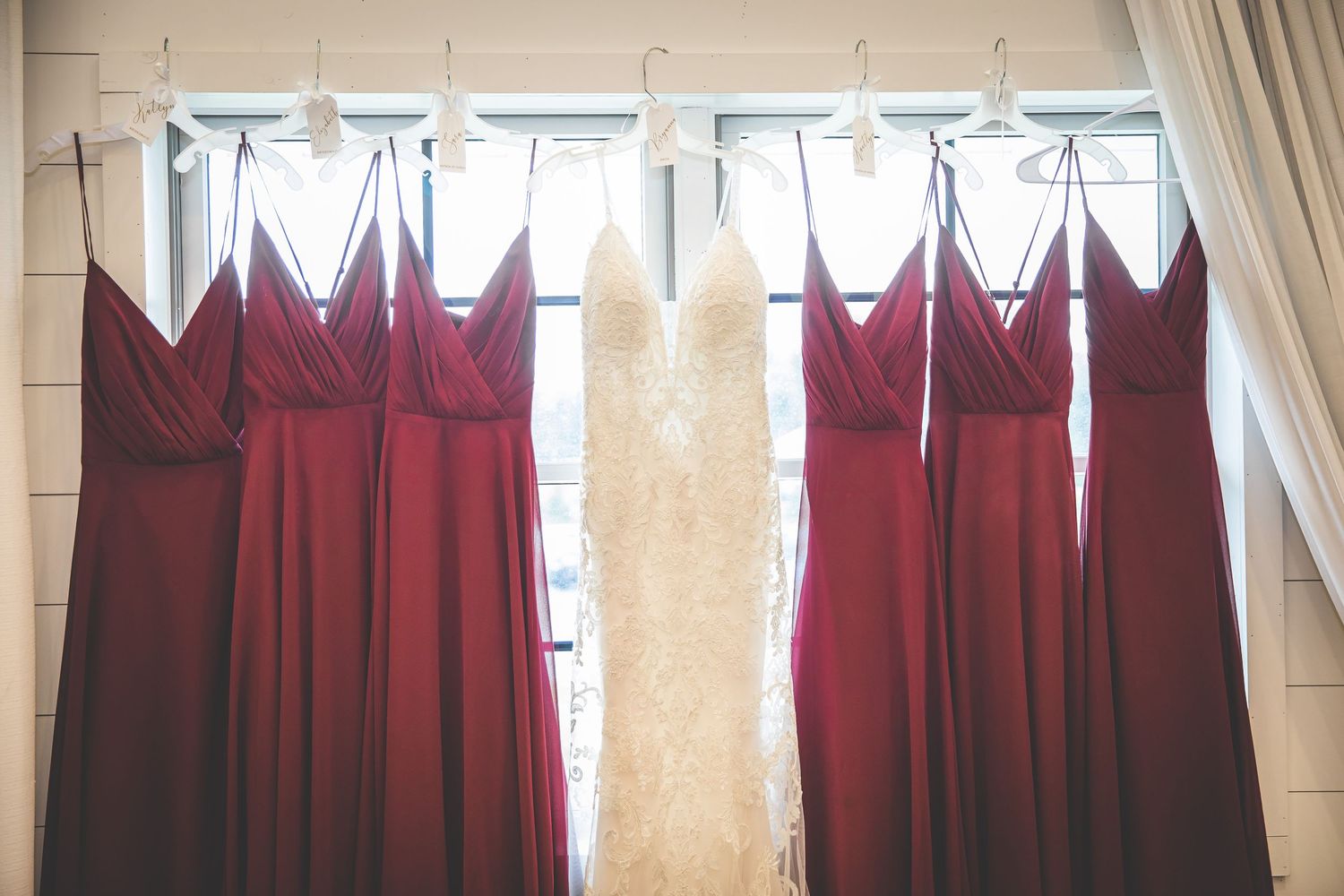 Row of burgundy bridesmaid dresses hanging by windows in preparation room.