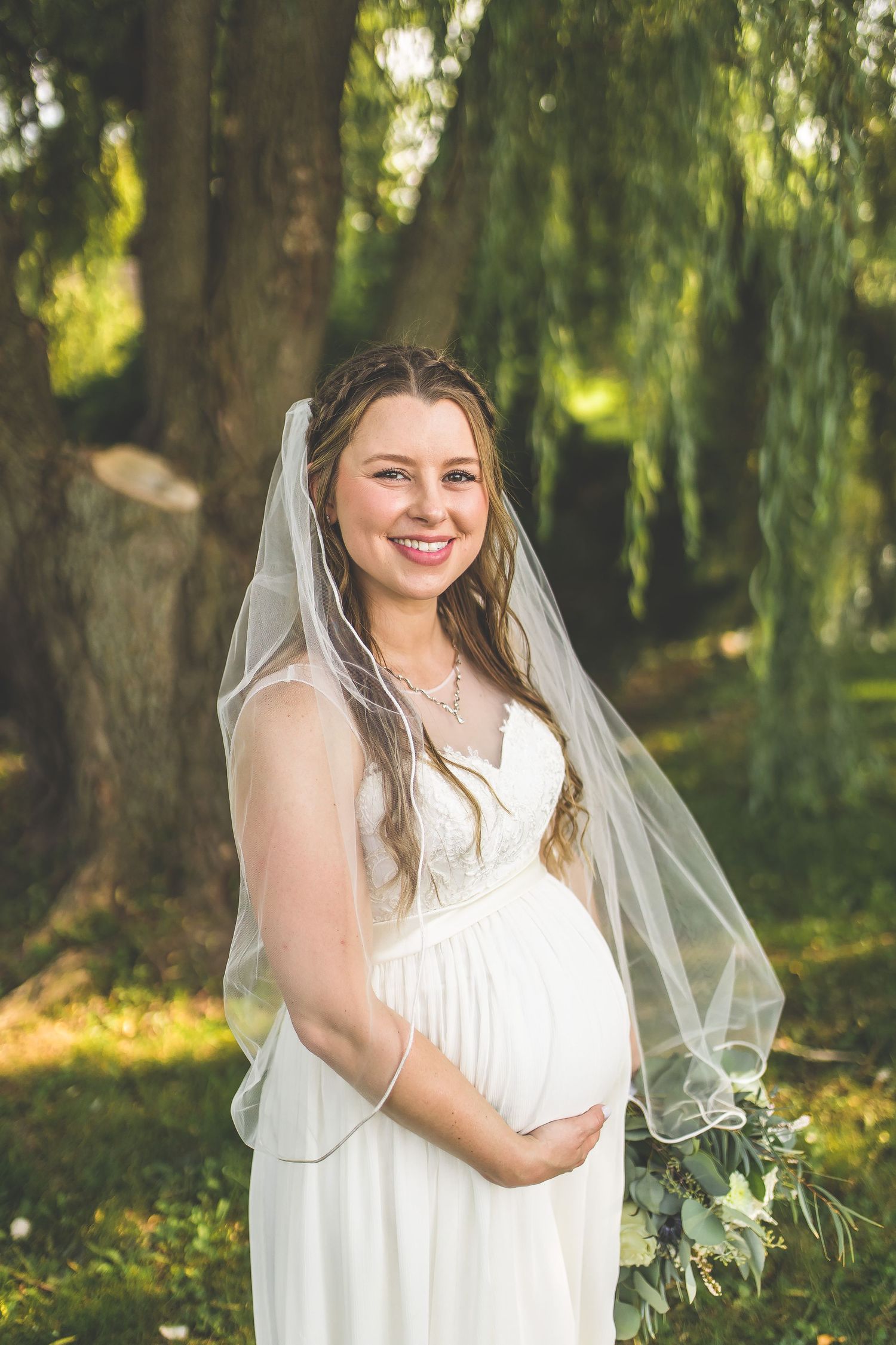 Bride in white dress holds elegant bouquet while posing under willow trees.