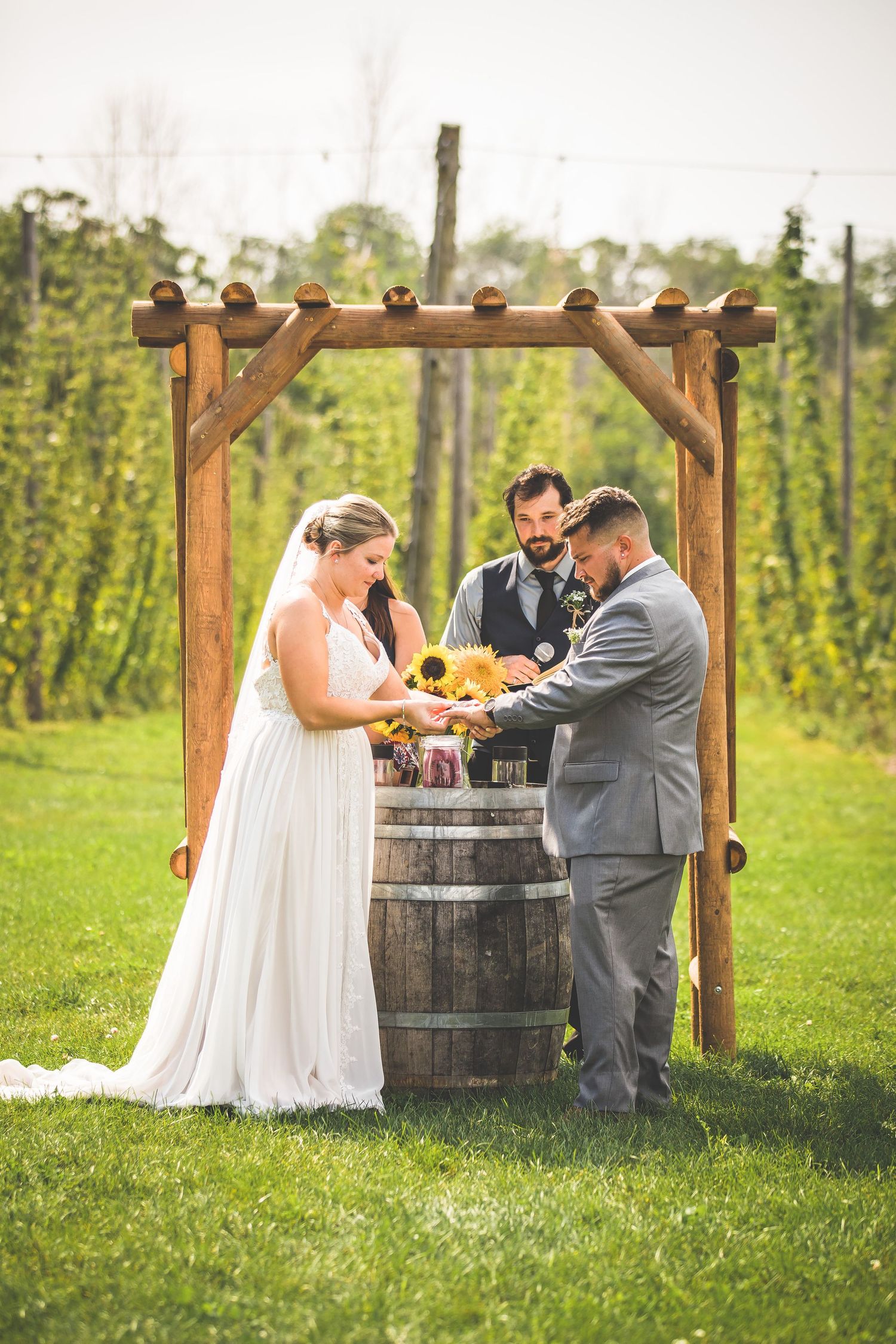 Outdoor wedding ceremony with wooden pergola and barrel altar in sunlit garden setting.