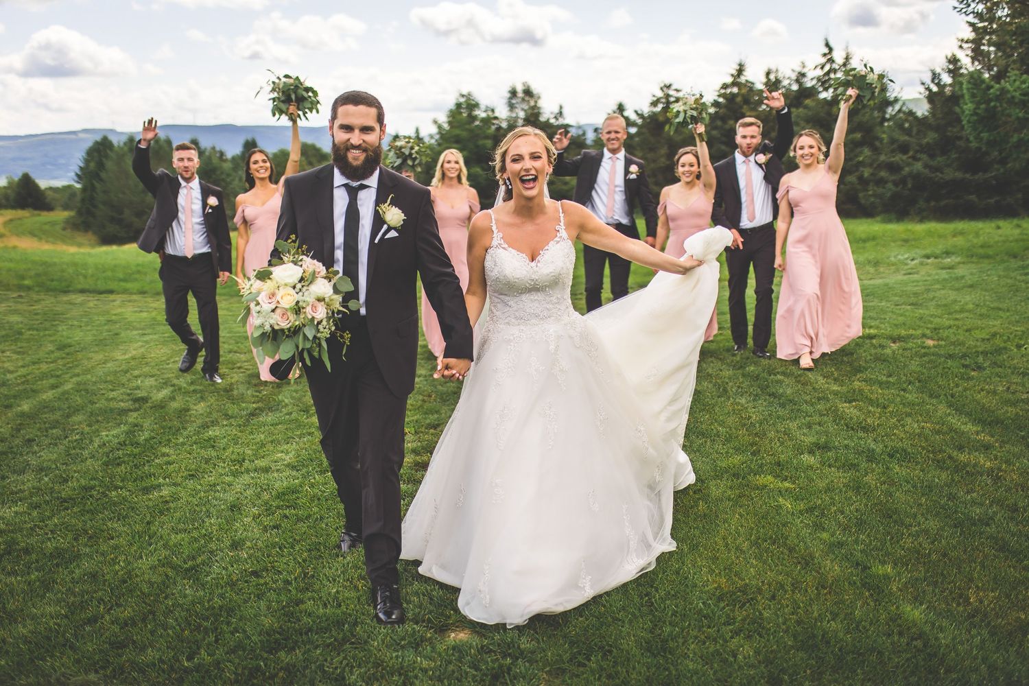 Wedding party poses casually on green lawn with mountain landscape in background.