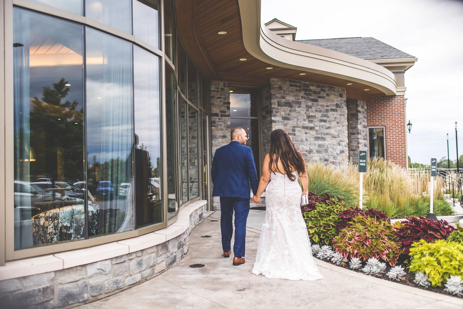 Couple walks along curved stone path outside modern wedding venue.