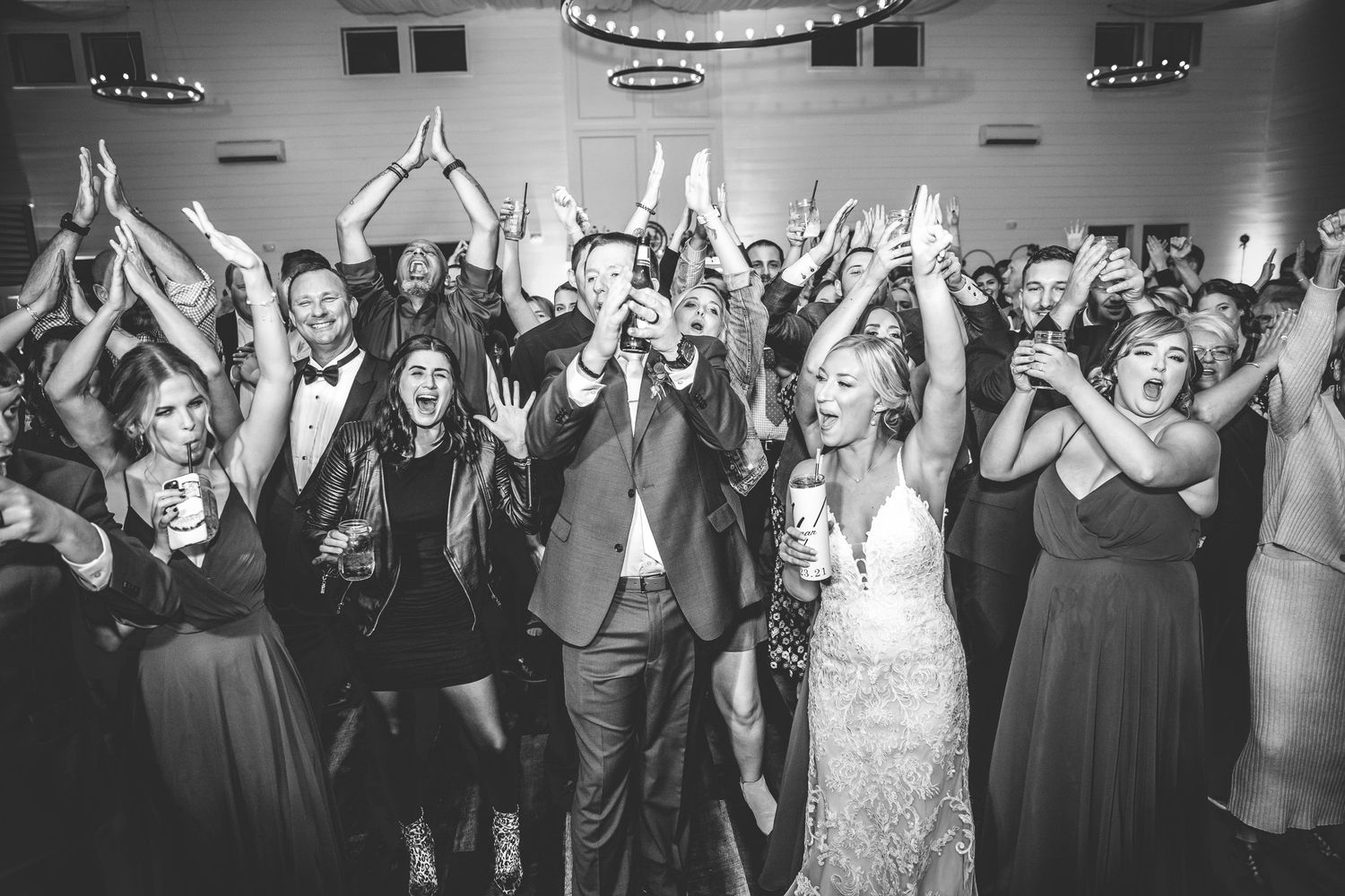 Black and white photo of wedding guests with raised arms celebrating on dance floor.