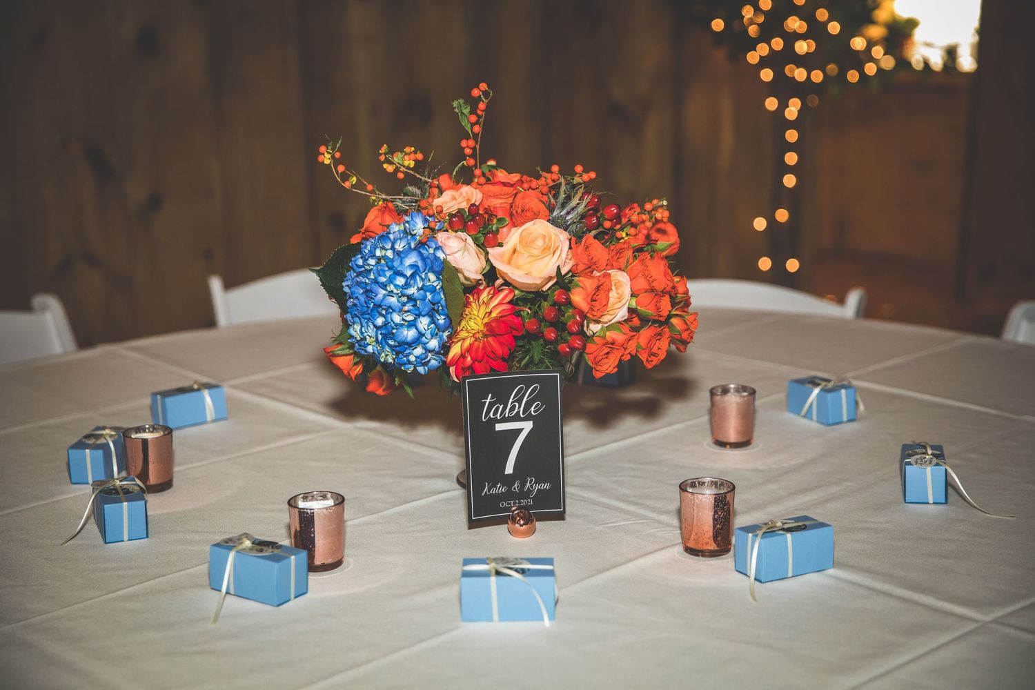 Wedding reception table decorated with blue and coral floral centerpieces and tealight candles in a rustic barn setting.