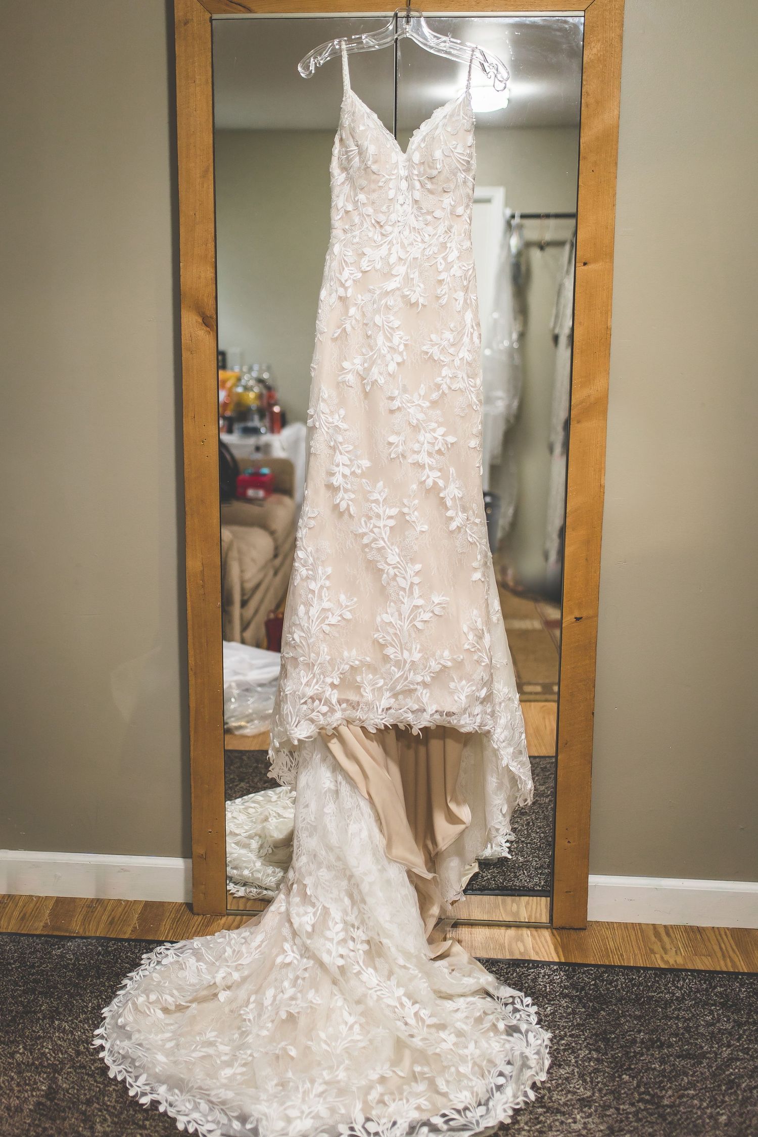 Elegant lace wedding dress with intricate floral details displayed on a wooden-framed mirror against a neutral wall.