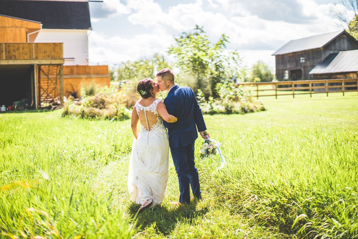 A couple walks together through a sunny green field near a rustic barn on their wedding day.