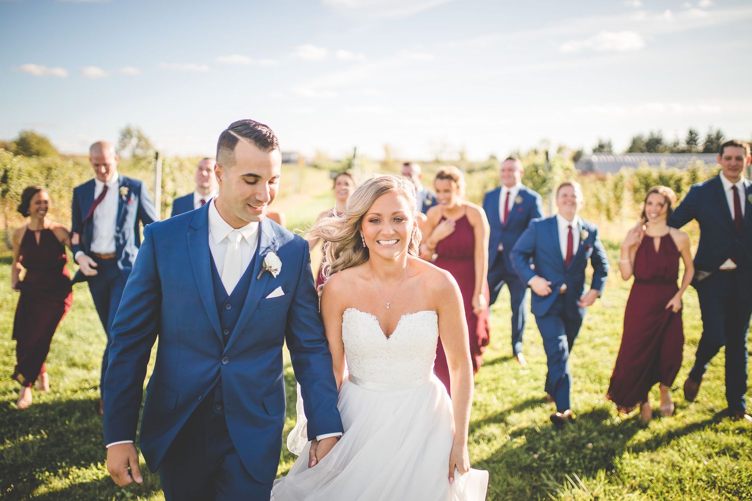 Wedding party in navy suits and burgundy dresses poses together in a sunny outdoor field.