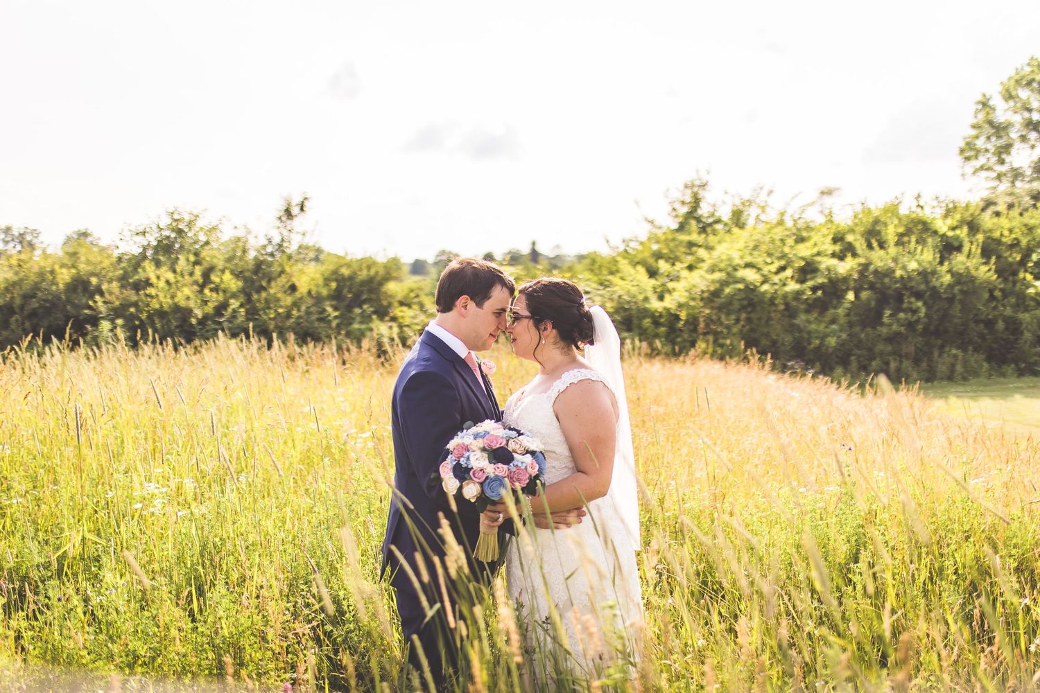 Romantic sunset portrait of newlyweds standing in tall grass holding a bouquet.