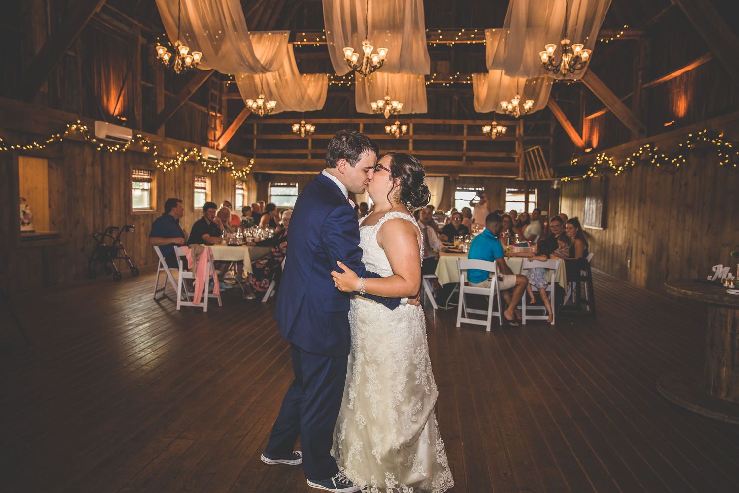 Couple shares their first dance in a rustic barn wedding venue decorated with twinkling lights and chandeliers.