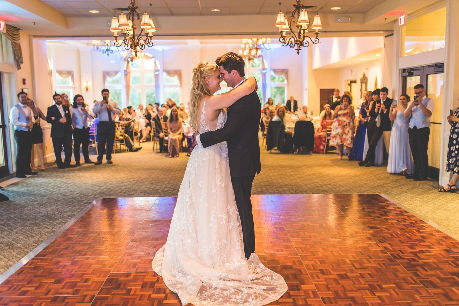 Wedding couple dances on a wooden floor surrounded by guests in an elegant reception venue.