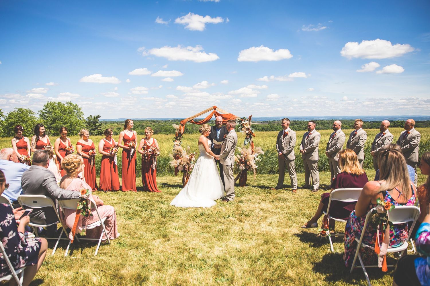 Outdoor wedding ceremony setup with white chairs on grassy hill under blue summer sky.