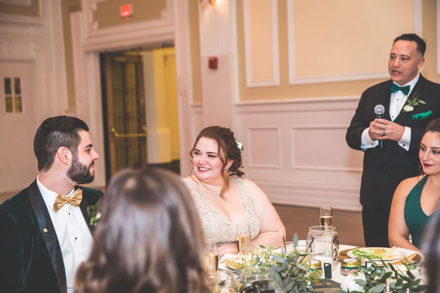 Wedding reception guests seated at decorated tables with floral centerpieces in a formal ballroom.