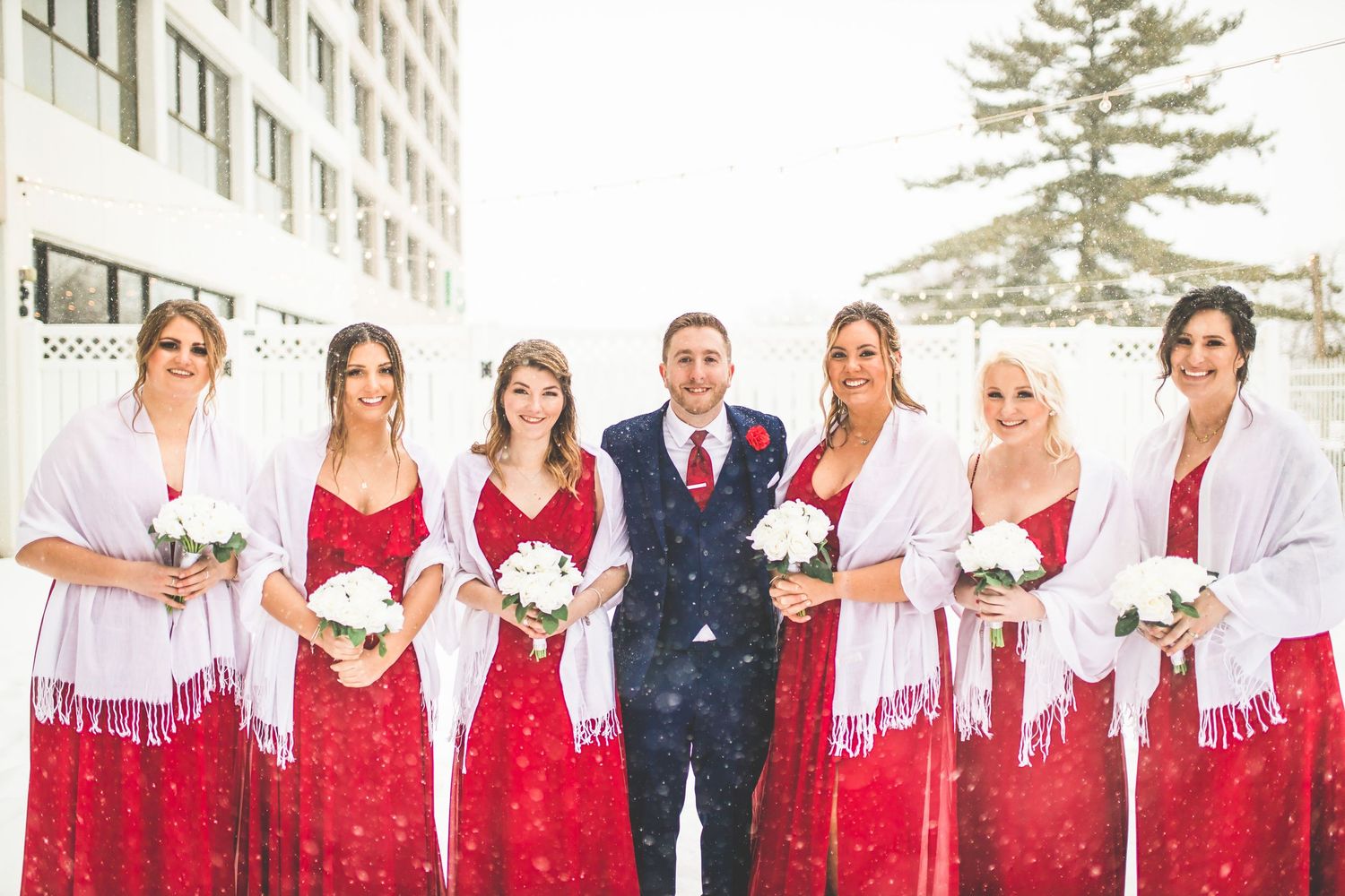 Wedding party members wear red dresses and white shawls while posing in the snow.