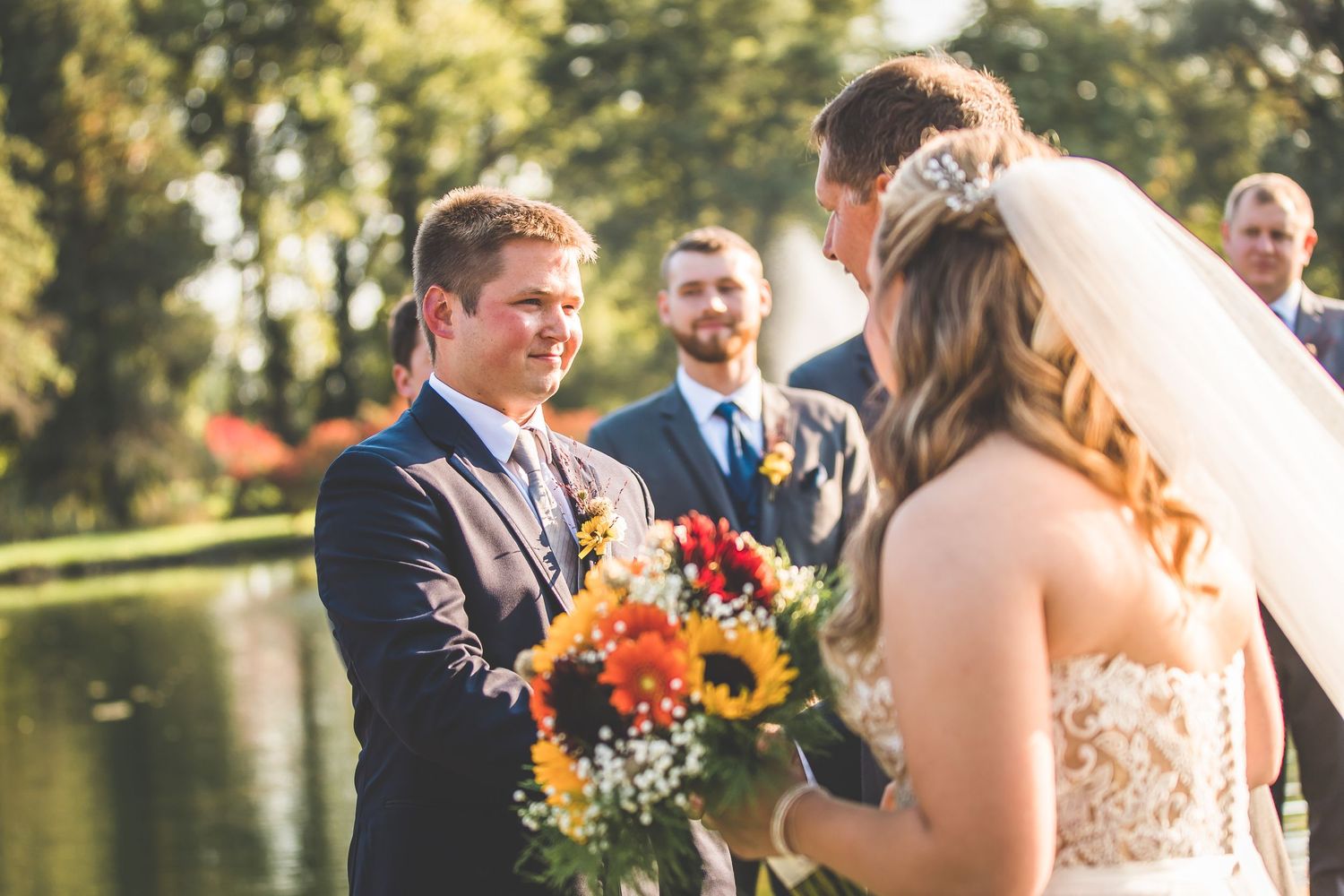 An outdoor wedding ceremony moment captured near a serene pond with sunflowers in the bouquet.
