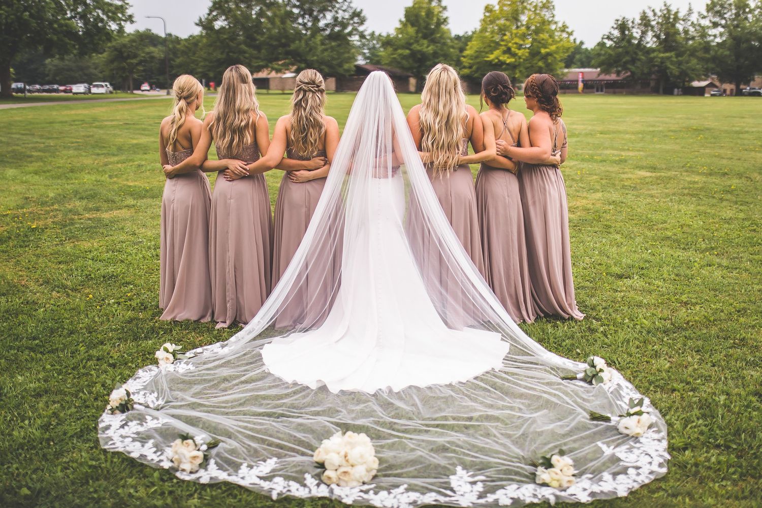 Bride's lace-trimmed cathedral veil spreads beautifully on grass surrounded by bridesmaids.