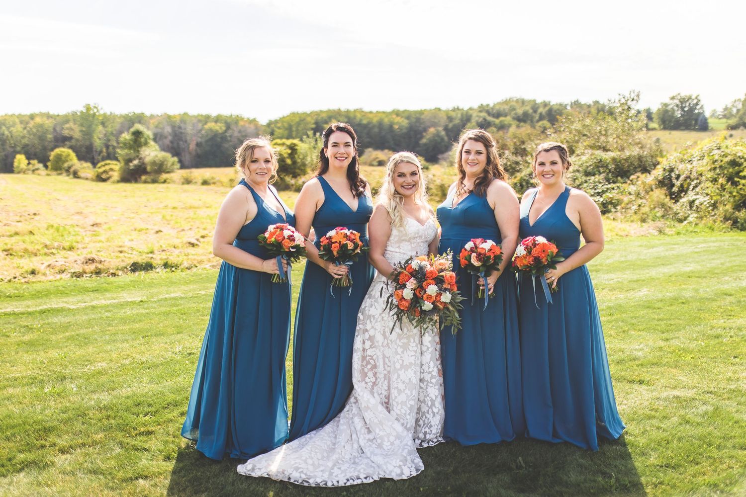 Bridal party wearing navy blue dresses and holding vibrant orange bouquets poses outdoors.