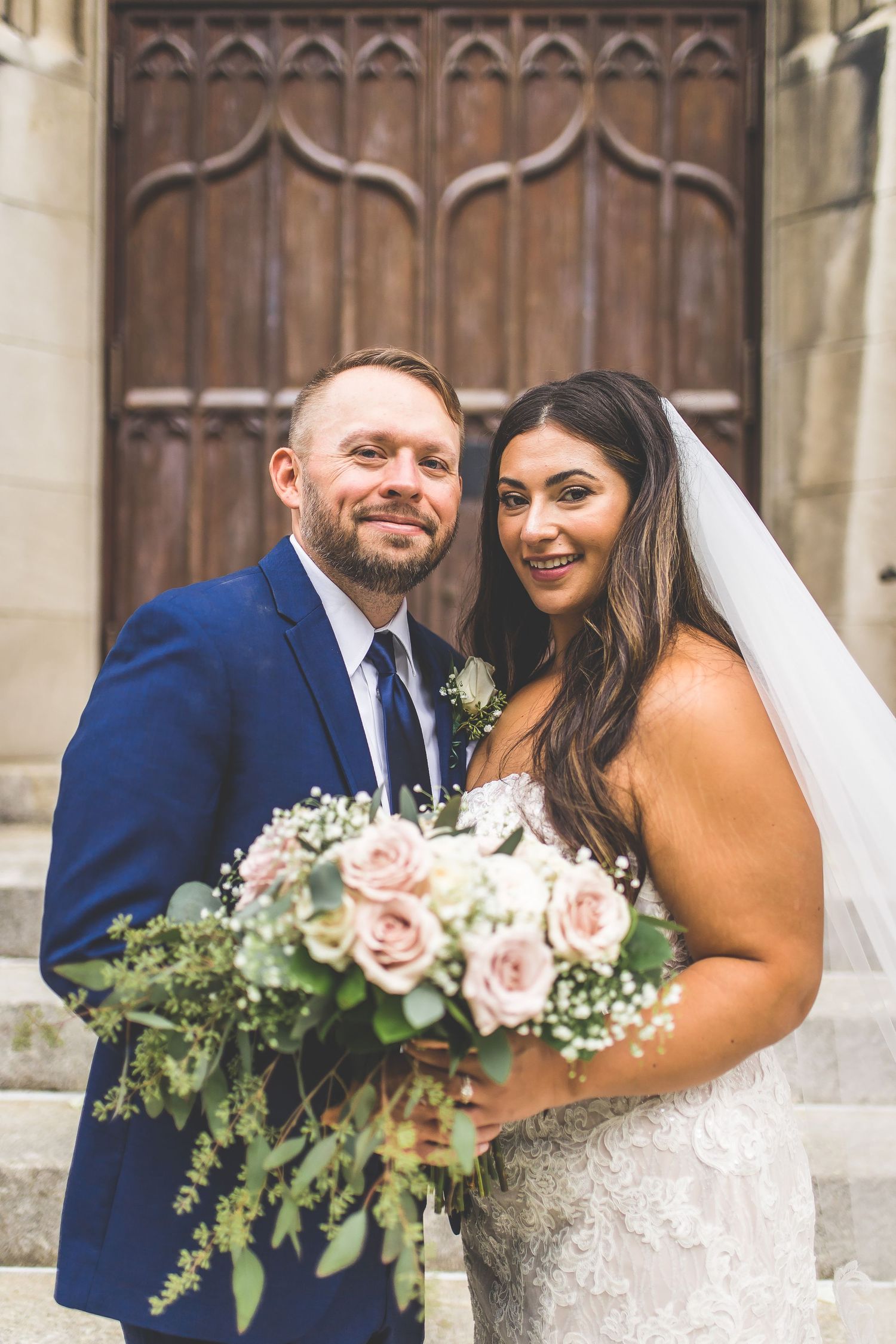Happy couple poses together outside historic church doors holding romantic pink and white bouquet.