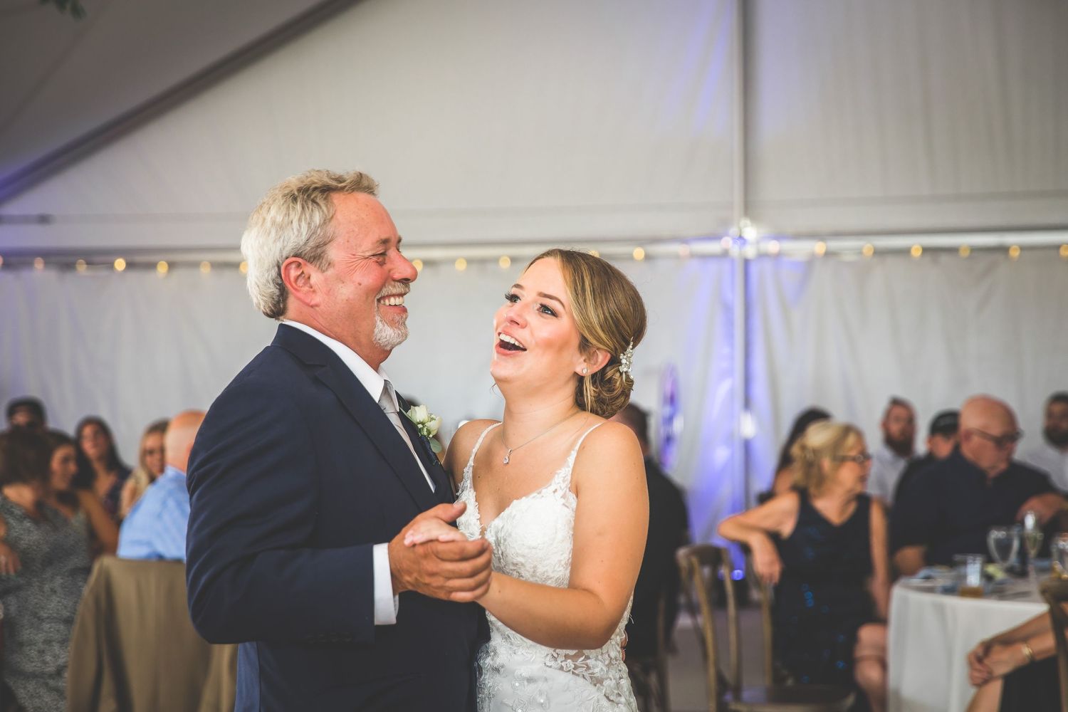 Wedding guests watch as a couple shares an emotional dance moment under string lights in a reception tent.