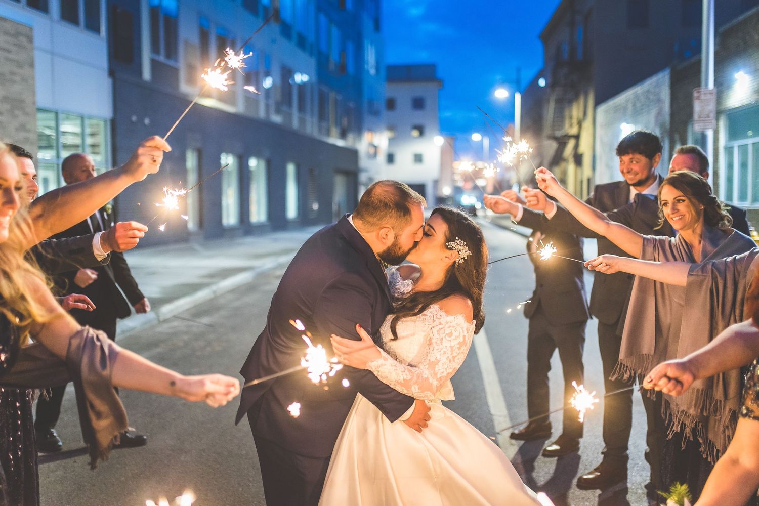 Wedding couple embraces on urban street while guests hold sparklers creating magical nighttime atmosphere.