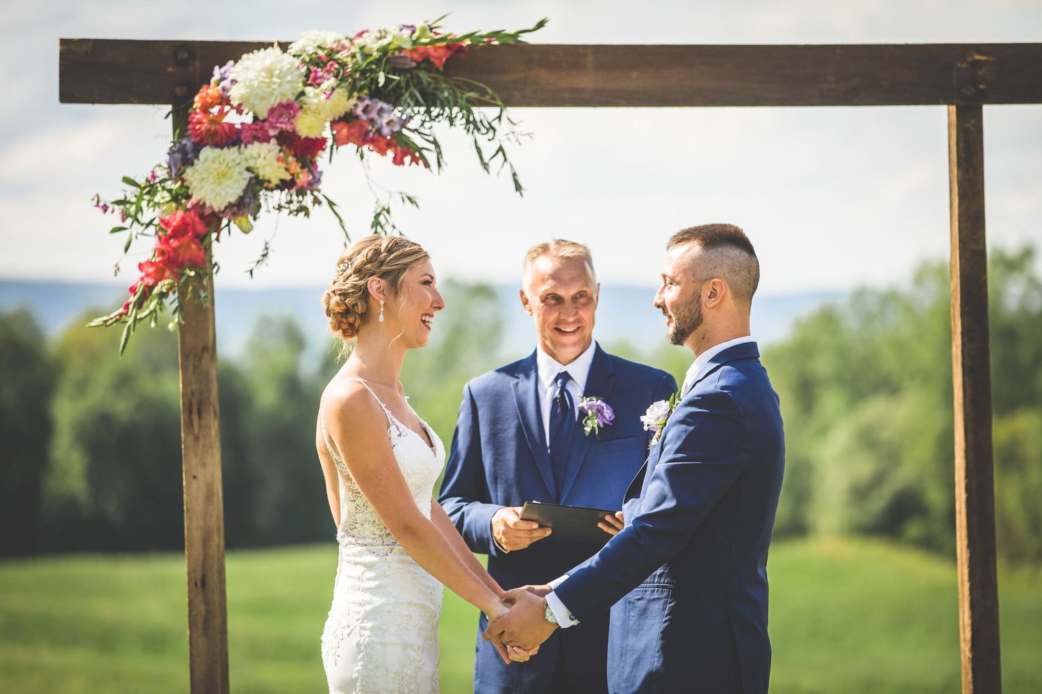 Wedding ceremony under wooden arbor decorated with pink flowers against scenic mountain backdrop.