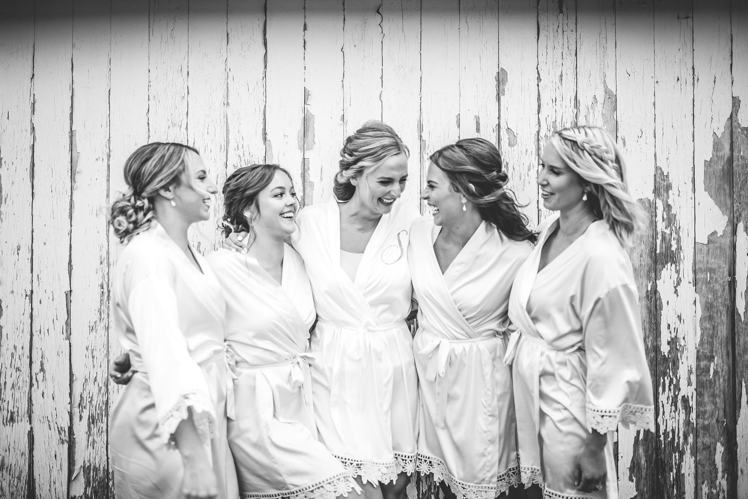 Wedding party in white robes sharing candid moments by rustic fence.