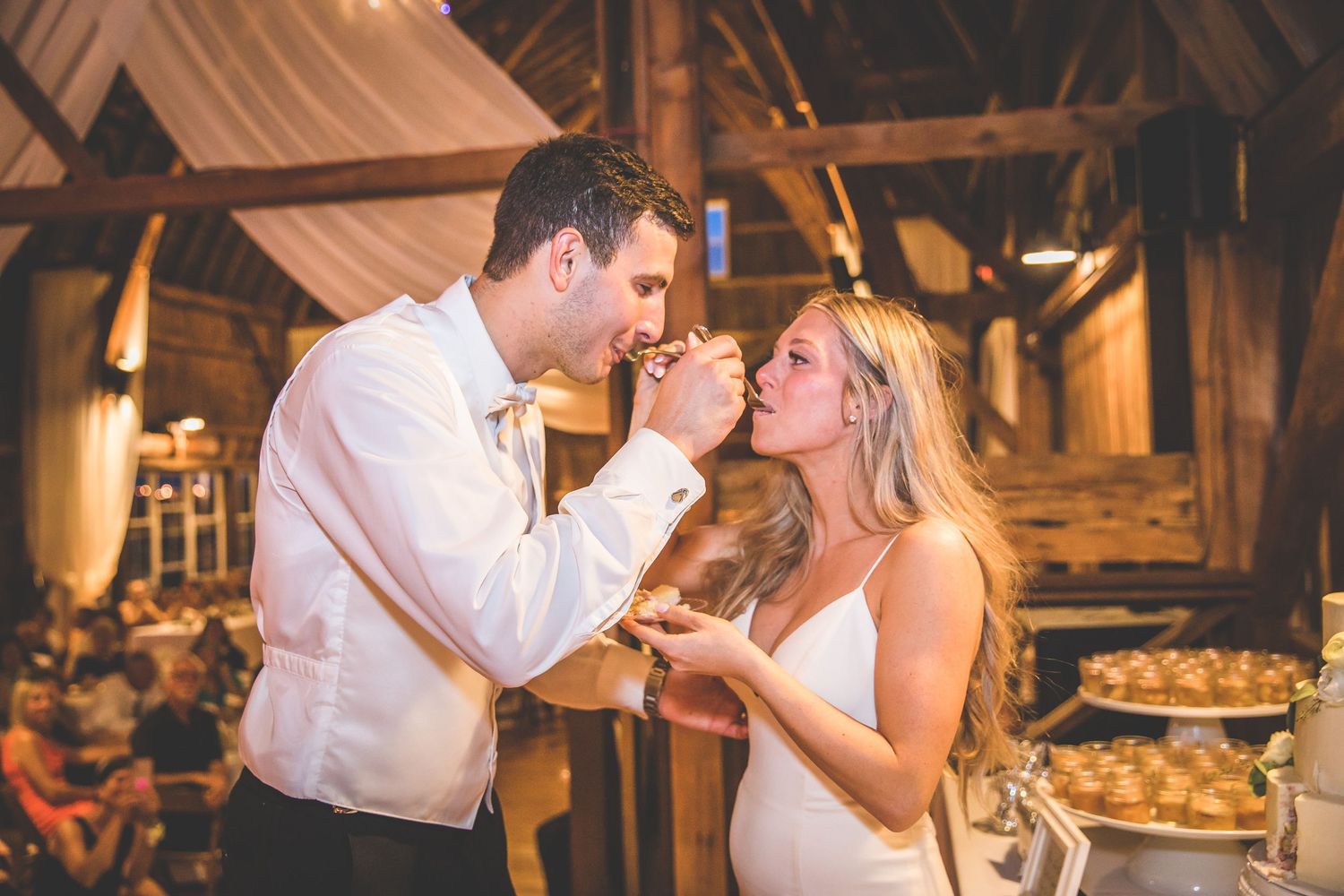 A couple shares a playful moment while feeding each other wedding cake at their rustic barn reception.