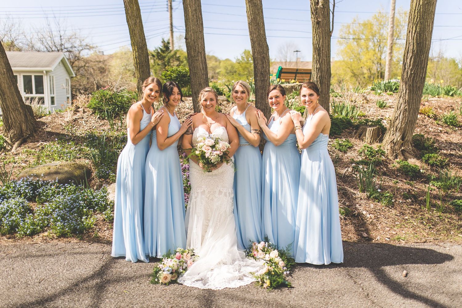 Wedding party in light blue dresses poses together outdoors among trees.