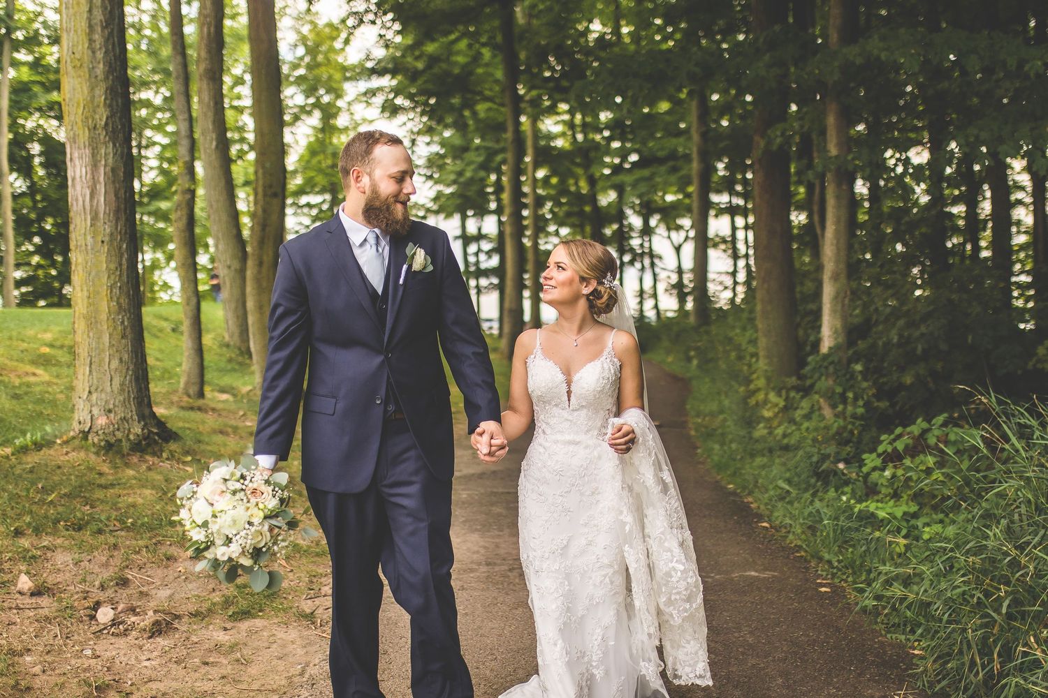 Newlyweds walking and embracing along a scenic wooded path during their wedding photoshoot.