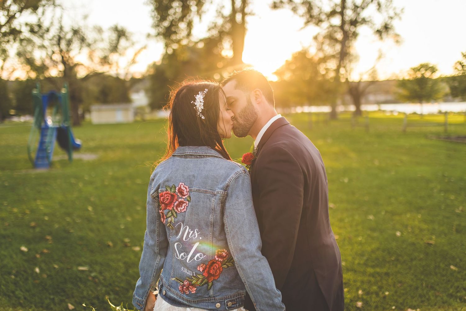 Bride wearing customized denim jacket poses with groom in sunlit field.