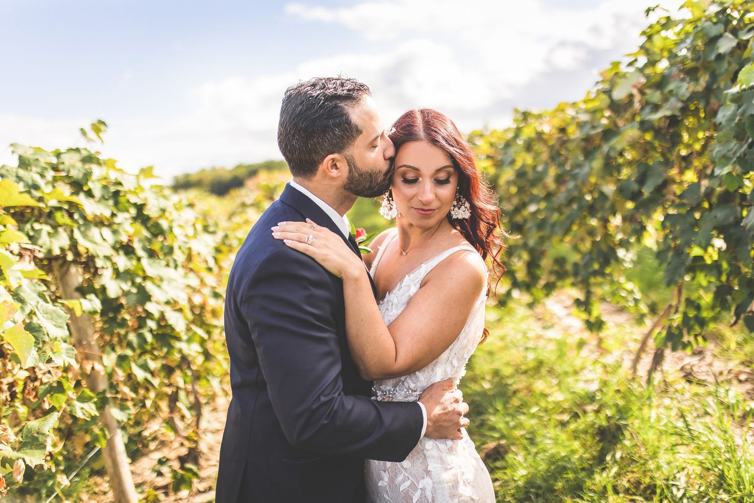 Wedding couple poses romantically between rows of grape vines on a sunny afternoon.