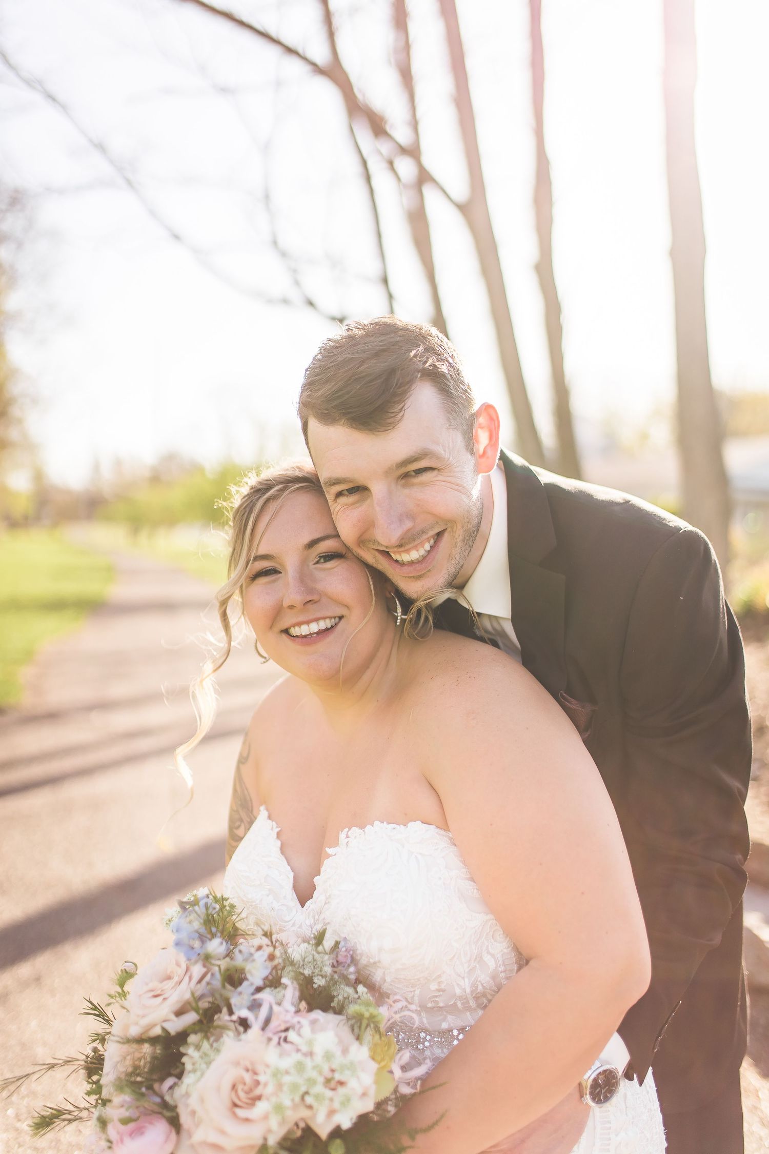 Joyful wedding couple embraces outdoors in natural sunlight with pink and white floral bouquet.