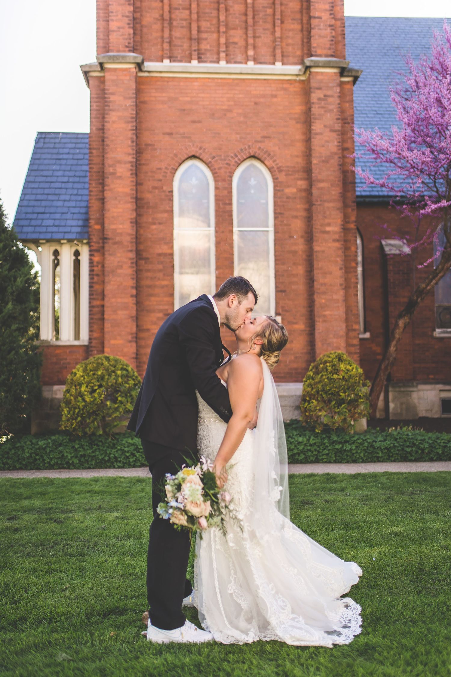Romantic wedding couple kisses outside red brick church with spring blossoms.