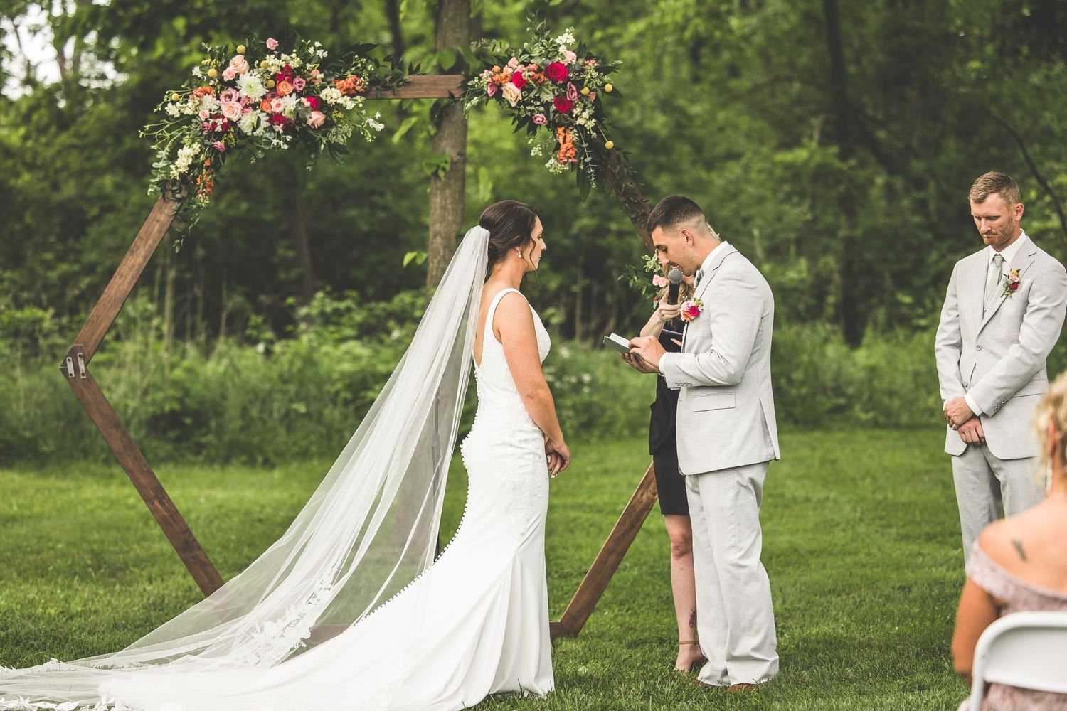 Outdoor wedding ceremony under wooden arch decorated with pink and coral flowers.