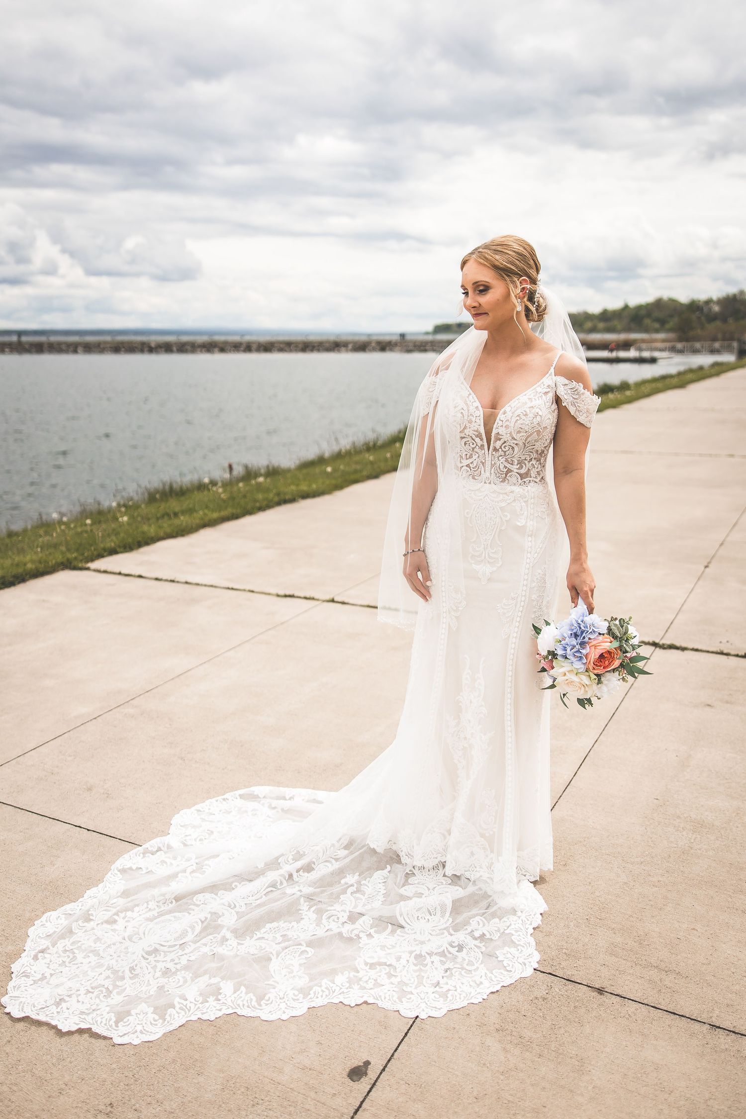 Elegant bride in lace wedding gown with long train stands on waterfront path holding colorful bouquet.