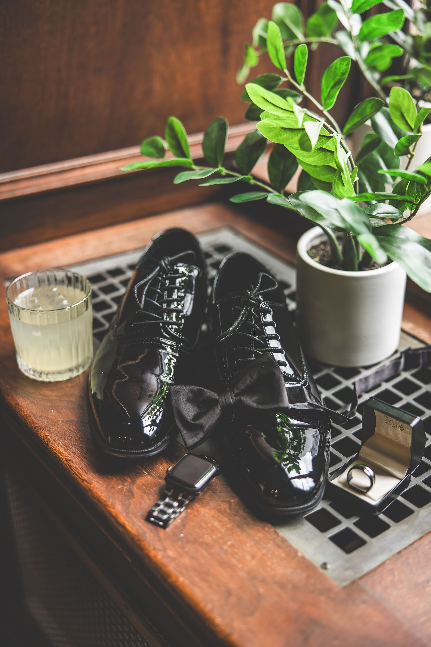 Shiny black dress shoes on laptop with houseplant and glass of water on wooden desk.