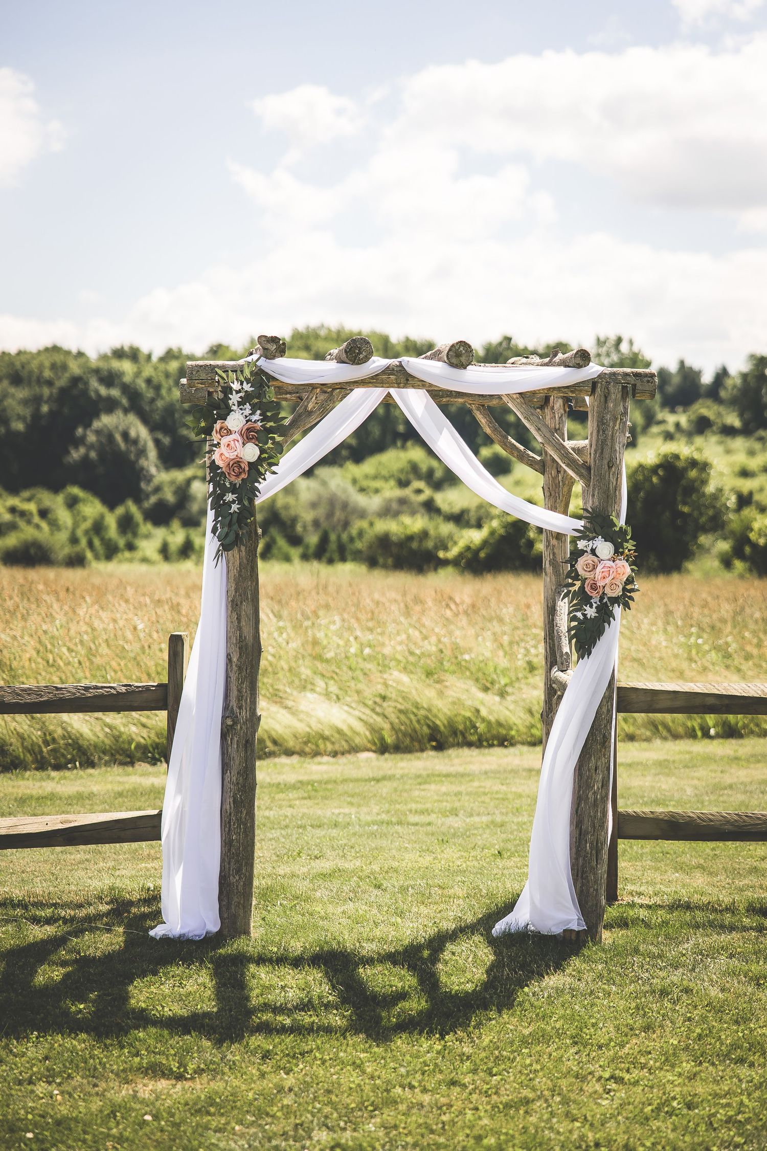 Rustic wedding arch decorated with white fabric and floral arrangements in an outdoor setting.