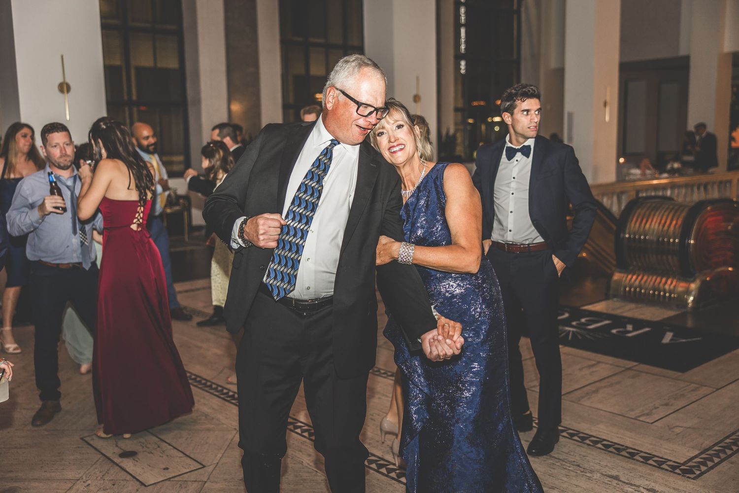 Wedding celebration with guests dancing in formal attire inside an elegant venue with columns.